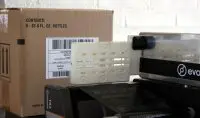 Cardboard box with a 'CONTENTS: 6 - 33.8 FL. OZ. BOTTLES' label beside a white plastic mounting attached to a printer-like device in a workshop setting.