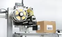 Automated packaging machine with a yellow reel and large metal wheel beside brown cardboard boxes on a conveyor.