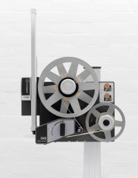 Industrial film projector with large metal reels on a vertical stand against a white brick wall